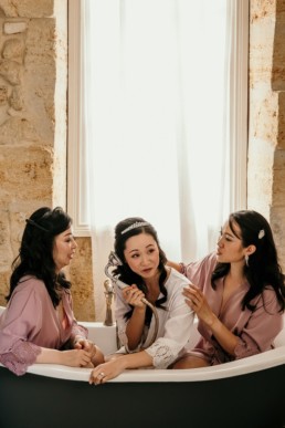 Bride and bridesmaids at wedding in chateau in Bordeaux, sitting in bath pretending to make call on shower