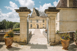 View through cream stone and wrought iron entrance gates across cobbled stone bridge to courtyard in front of the Aristocratic chateau wedding venue in the Dordogne
