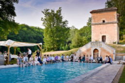 Wedding guests seated around pool with feet in the water with stone tower and trees behind at theAristocratic chateau wedding venue in the Dordogne