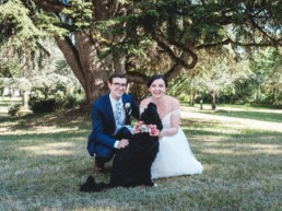 Bride and groom sitting with their large black dog who is wearing a floral collar
