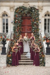 Bride and 6 bridesmaids in two lines dressed in claret coloured long dresses with autumnal bouquets standing on steps of chateau with huge autumnal floral arch over door behind and flowers up the steps