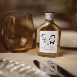 Amber water glass on table next to miniature bottle of vodka with label with caricature of bride and groom